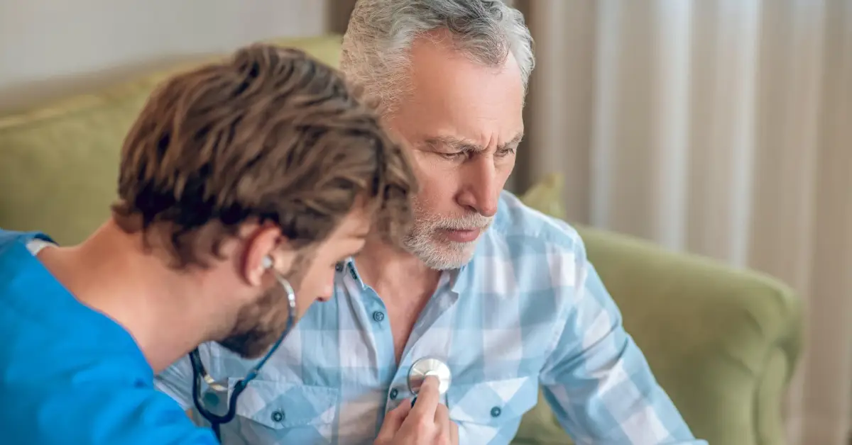 A nurse in blue scrubs using a stethoscope to listen to an older adult seated on a couch at home